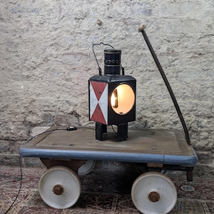 Op de afbeelding: Een vintage zwart metalen spoorweglantaarn met een rood-wit geblokt zijpaneel, verlicht door een interne lamp, rustend op een houten industriële kar met vier grote witte wielen en een lange metalen hendel die omhoog steekt.