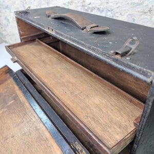 May include: A rectangular black wooden tool box with a weathered finish, featuring a brown leather handle on top, metal latches, and a hinged lid that opens to reveal an interior wooden tray and a natural wood grain base inside the main compartment.