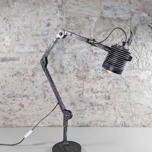 Könnte beinhalten: Industrielle Schreibtischlampe mit dunkelgrauer Oberfläche. Die Lampe hat einen beweglichen Arm und einen zylindrischen Lampenkopf. Der Sockel ist rund, und die Lampe ist an eine Steckdose angeschlossen. Der Hintergrund ist eine strukturierte Backsteinmauer.