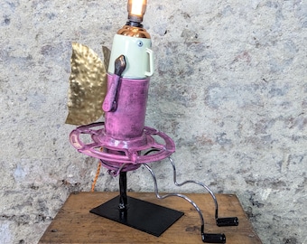 May include: A whimsical, handmade lamp sculpture. It features a light bulb atop a repurposed metal and ceramic body, with a pink base and a gold-colored wing. The lamp is mounted on a black base and has wire legs.