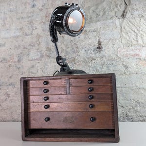 Może przedstawiać: Lampa biurkowa w stylu industrialnym z czarnym metalowym kloszem i regulowanym ramieniem, zamontowana nad zabytkową drewnianą skrzynką narzędziową z wieloma szufladami i czarnymi gałkami. Lampa jest włączona.