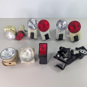 May include: A selection of vintage bicycle lights and reflectors. The lights feature clear or red lenses, and are set in beige or black casings. Some are round, others rectangular. Several black mounting brackets are also present.