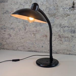 Op de afbeelding: Zwarte bureaulamp met een gebogen, verstelbare nek en een ronde voet. De lamp heeft een zwarte lampenkap met een gloeilamp erin. De lamp is aangesloten op een stopcontact met een zwart snoer.