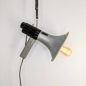 May include: A vintage-style megaphone light fitting with a black and silver finish. The light is hanging from a black leather strap and has a clear light bulb.
