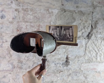 Op de afbeelding: Een antieke stereoscoop viewer van metaal en hout. De viewer heeft een donker, gebogen oculair en een houten handvat. Een vintage foto is in de viewer geplaatst. De achtergrond is een getextureerde, lichtgeleurde bakstenen muur.