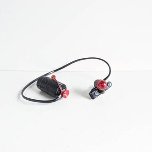 May include: A black bicycle light set with a red lens and a black cylindrical battery holder. The set includes a black wire connecting the two components, and a red fastener on the battery holder.