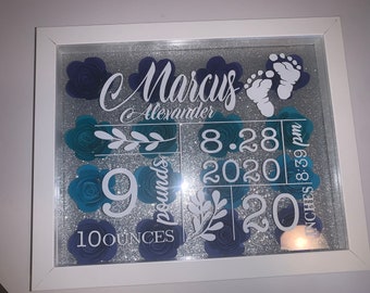 May include: A white framed picture with a silver glitter background. The picture is decorated with blue paper flowers and white lettering. The lettering says "C Marcus C Alexander", "9 pounds 100 ounces", "8.28.2020 8:59 pm", and "20 inches".