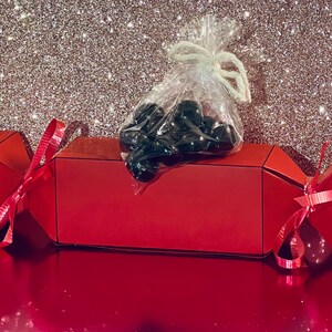 Puede incluir: Un petardo navideño rojo brillante con lazos rosas y una bolsa transparente de caramelos negros encima. El petardo está sobre una superficie roja brillante con un fondo brillante.