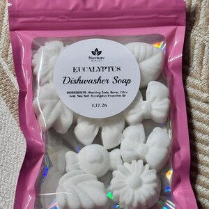 Op de afbeelding: Een roze zak met ritssluiting gevuld met witte vaatwasmiddelzeep in de vorm van bloemen en strikken. De zak heeft een doorzichtig venster met de tekst "EUCALYPTUS Dishwasher Soap" op een wit etiket.