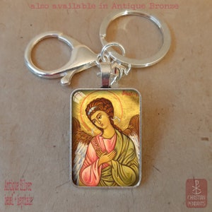 Puede incluir: Llavero con bisel plateado antiguo con un colgante rectangular con una ilustraci&oacute;n de un &aacute;ngel. El &aacute;ngel tiene cabello casta&ntilde;o, un halo y viste t&uacute;nicas rosas y verdes. El texto "also available in Antique Bronze" es visible.