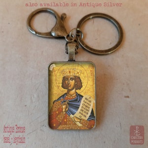 May include: Antique bronze keychain with a rectangular pendant featuring a religious icon. The icon depicts a figure with a crown, holding a scroll. Text on the image reads "also available in Antique Silver" and "Antique Bronze bezel + keychain."