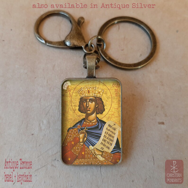 May include: Antique bronze keychain with a rectangular pendant featuring a religious icon. The icon depicts a figure with a crown, holding a scroll. Text on the image reads "also available in Antique Silver" and "Antique Bronze bezel + keychain."