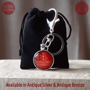 May include: A keychain with a round, red pendant featuring a Christian symbol and the words "CHRISTIAN PENDANTS" in gold. The keychain is attached to a silver clasp and key ring, and is next to a black velvet bag. The text "Available in Antique Silver & Antique Bronze" is at the bottom.