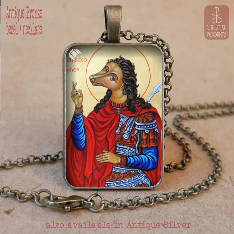 Puede incluir: Collar con colgante de bronce antiguo. El colgante rectangular presenta una imagen de San Crist&oacute;bal con cabeza de perro sobre un fondo dorado. Cuelga de una cadena a juego. La palabra "CHRISTOPHER" es visible.