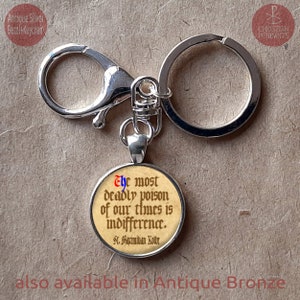 May include: Antique silver bezel keychain with a round pendant. The pendant features the quote "The most deadly poison of our times is indifference" attributed to St. Maximilian Kolbe. Also available in Antique Bronze.
