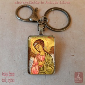 Puede incluir: Llavero de bronce antiguo con un colgante rectangular que presenta un icono religioso de un &aacute;ngel. El &aacute;ngel tiene cabello casta&ntilde;o, un halo dorado y viste t&uacute;nicas rosas y verdes. El texto "also available in Antique Silver" es visible.