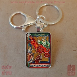Pode incluir: Porta-chaves prateado com um pendente retangular com uma ilustra&ccedil;&atilde;o religiosa. A obra de arte retrata figuras, uma carro&ccedil;a vermelha e um cavalo. O texto "also available in Antique Bronze" &eacute; vis&iacute;vel.