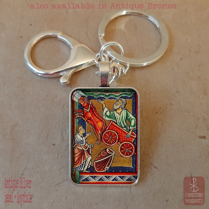 Pode incluir: Porta-chaves prateado com um pendente retangular com uma ilustra&ccedil;&atilde;o religiosa. A obra de arte retrata figuras, uma carro&ccedil;a vermelha e um cavalo. O texto "also available in Antique Bronze" &eacute; vis&iacute;vel.
