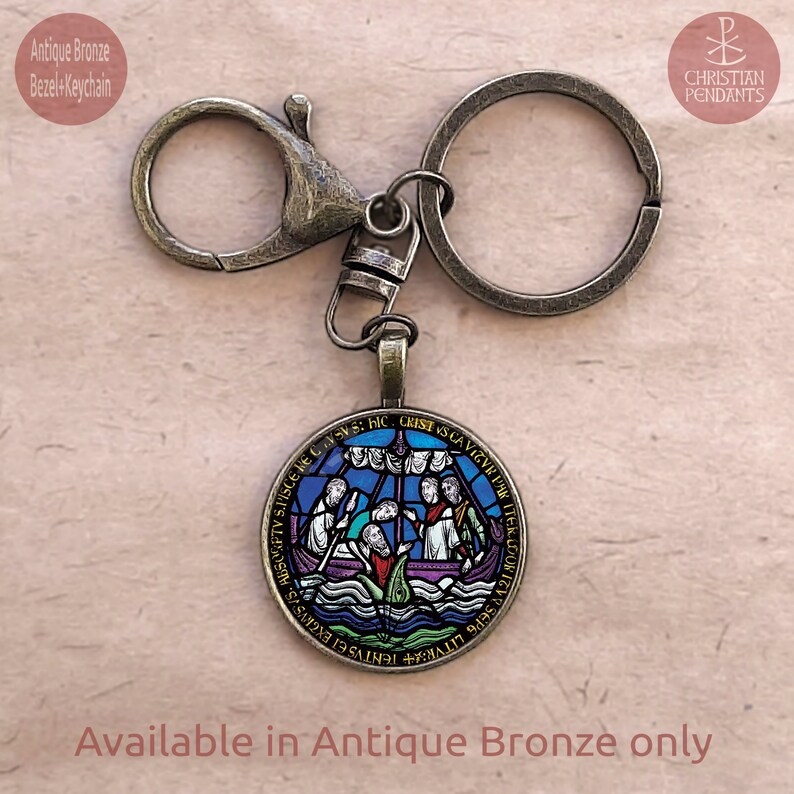 May include: Antique bronze keychain with a round pendant depicting a stained-glass boat scene. The pendant features blue, purple, and green hues. The keychain includes a lobster clasp and a key ring. The text "Available in Antique Bronze only" is visible. The measurements are in cm.