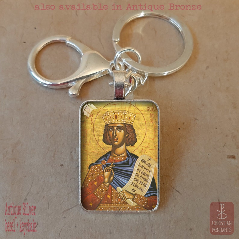 May include: Silver keychain with a rectangular pendant featuring a religious icon. The icon depicts a figure with a crown, holding a scroll. The background is gold with text. The keychain has a lobster clasp and a split ring.