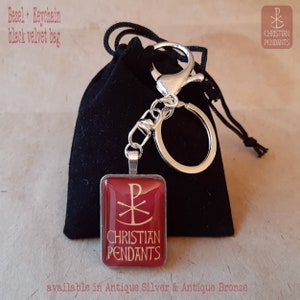 May include: A red and silver keychain pendant with the text "CHRISTIAN PENDANTS" and a religious symbol. The pendant is attached to a silver keychain and is presented with a black velvet bag. The text "Bezel + Keychain black velvet bag" is visible.