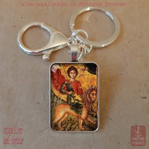 May include: Antique silver bezel keychain featuring a religious icon. The rectangular pendant displays a detailed image of a figure riding a lion, with text that reads "Christian Pendants". The keychain also has the text "also available in Antique Bronze".