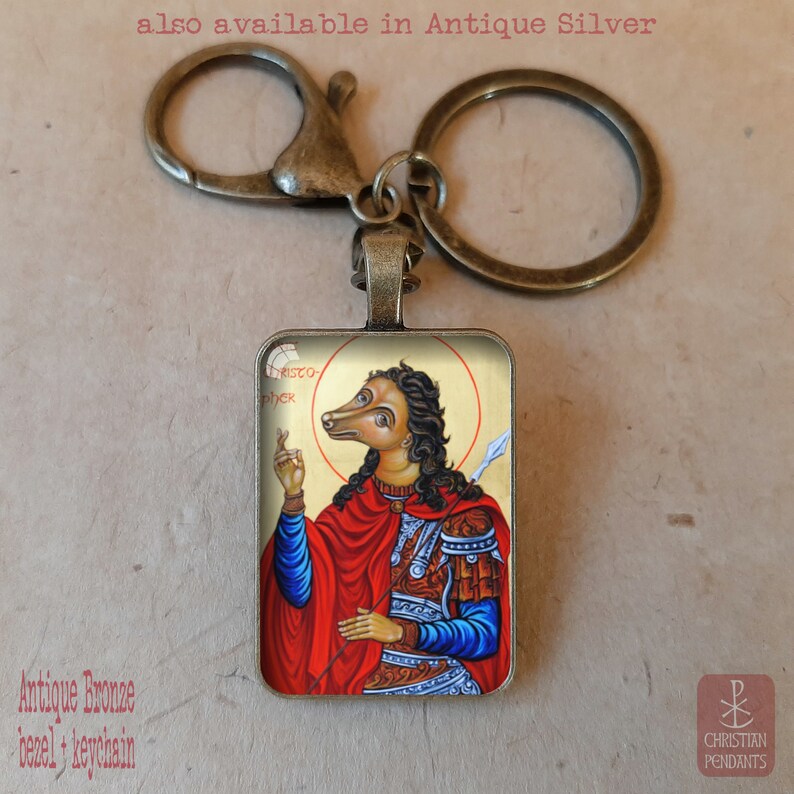 Puede incluir: Llavero de bronce antiguo con un colgante rectangular que muestra un icono religioso de una figura con cabeza de perro en rojo y azul. El texto "also available in Antique Silver" es visible en la parte superior. Las palabras "Antique Bronze bezel + keychain" est&aacute;n en la parte inferior.