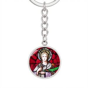 May include: Silver keychain with a round glass charm featuring a stained glass image of a woman holding a chalice and a palm branch. The image is set against a red background with gold accents.