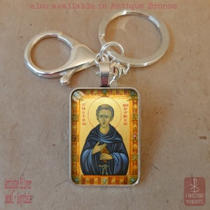 May include: A silver rectangular keychain featuring a religious icon. The icon depicts a man in blue robes with a gold halo, with the text "St. Palladius" and "Bishop of Ireland". The keychain includes a lobster clasp and a key ring. The text "also available in Antique Bronze" is visible.