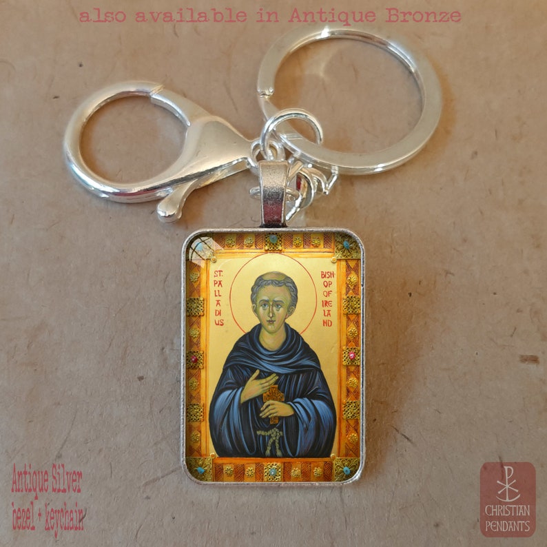 May include: A silver rectangular keychain featuring a religious icon. The icon depicts a man in blue robes with a gold halo, with the text "St. Palladius" and "Bishop of Ireland". The keychain includes a lobster clasp and a key ring. The text "also available in Antique Bronze" is visible.