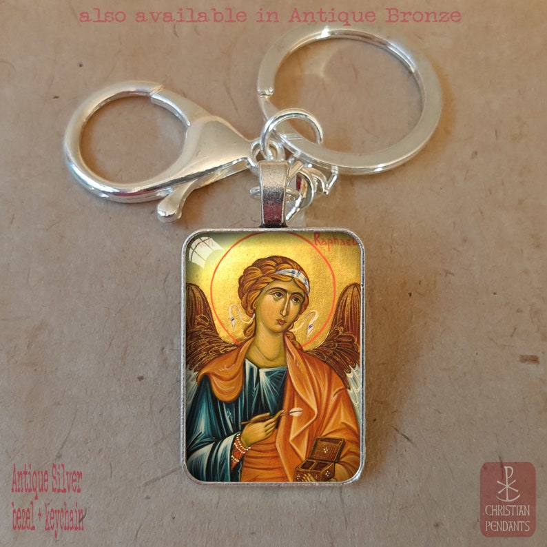 May include: Antique silver keychain with a rectangular pendant featuring an icon of an angel with a gold halo. The angel wears a blue robe and orange cloak, holding a small box and a stylus. Text on the image reads "Raphael" and "also available in Antique Bronze".