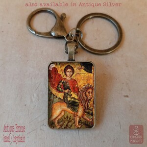 May include: Antique bronze keychain with a rectangular pendant featuring a religious icon of a figure riding a lion. The image has a vintage aesthetic. The text "also available in Antique Silver" is visible.