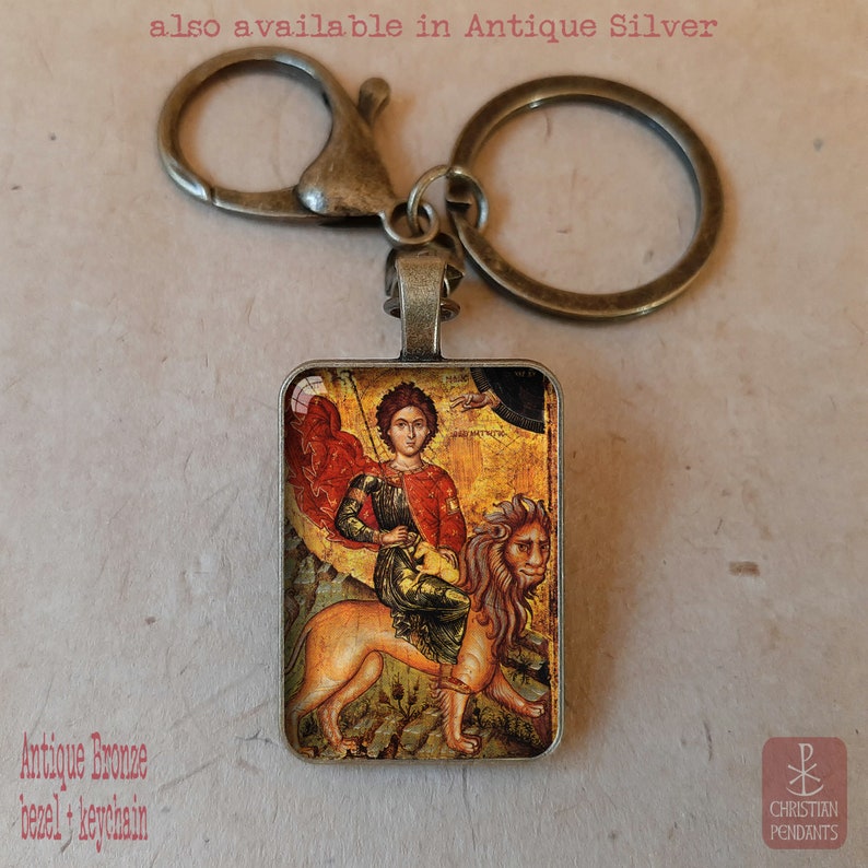 May include: Antique bronze keychain with a rectangular pendant featuring a religious icon of a figure riding a lion. The image has a vintage aesthetic. The text "also available in Antique Silver" is visible.