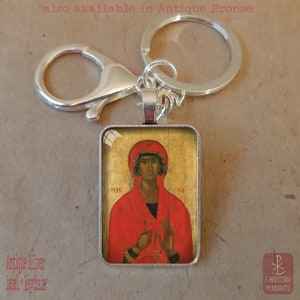 Puede incluir: Llavero con bisel plateado antiguo con un icono religioso. El colgante rectangular muestra una figura con t&uacute;nica y tocado rojos sobre un fondo dorado. El llavero incluye un mosquet&oacute;n y una anilla partida.