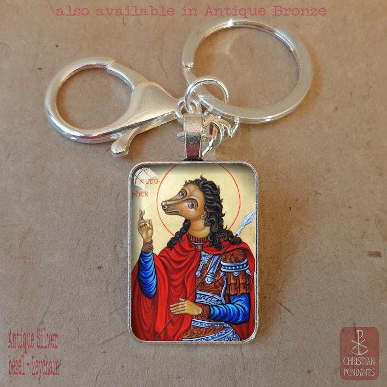 Puede incluir: Llavero con bisel plateado antiguo con un icono religioso de una figura con cabeza de perro vestida de rojo y azul, sosteniendo una lanza. La imagen tiene un fondo dorado y est&aacute; etiquetada como "Christo-pher". Tambi&eacute;n disponible en bronce antiguo.