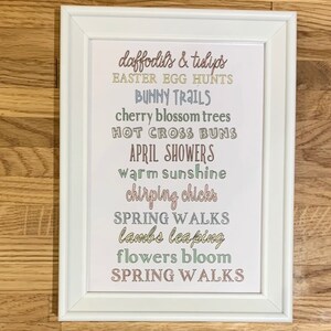 May include: A white framed print with a list of spring-themed words in different colours, including "daffodils & tulips", "Easter Egg Hunts", "Bunny Trails", "cherry blossom trees", "Hot Cross Buns", "April Showers", "warm sunshine", "chirping chicks", "Spring Walks", "lambs leaping", "flowers bloom", and "Spring Walks".