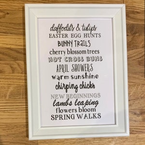 May include: A white framed print with black text listing spring-themed words and phrases, such as "daffodils & tulips", "Easter egg hunts", "bunny trails", "cherry blossom trees", "hot cross buns", "April showers", "warm sunshine", "chirping chicks", "new beginnings", "lambs leaping", "flowers bloom", and "spring walks".