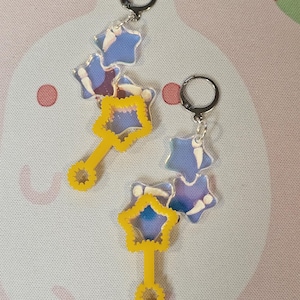 Puede incluir: Dos pares de pendientes con temática celestial. Cada pendiente presenta un aro plateado, conectado a un grupo de dijes en forma de estrella azul y morada. Debajo, cuelga un dije amarillo en forma de estrella con un asa. Los pendientes están sobre un fondo rosa claro.