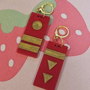Puede incluir: Un par de pendientes rectangulares rojos con detalles dorados. Un pendiente presenta un círculo dorado, mientras que el otro tiene dos triángulos dorados. Ambos pendientes tienen cierres dorados.