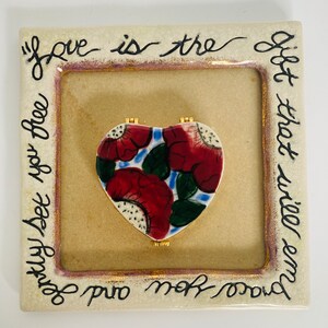 Puede incluir: Una placa de cerámica con una caja en forma de corazón en el centro. La caja está decorada con diseños florales rojos y verdes. La placa tiene un fondo blanco con letras negras que dicen "El amor es el regalo que te liberará y te fortalecerá".