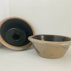 Puede incluir: Dos cuencos de cerámica con un esmalte marrón oscuro en el interior y un esmalte marrón claro en el exterior. El cuenco más grande tiene un diseño azul y blanco y un sello que dice "The Pottery Place, 1990".