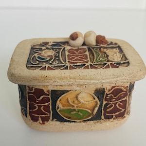 May include: A ceramic trinket box with a lid featuring a geometric design and two small white knobs. The box is decorated with brown, red, green, and black patterns.