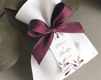 May include: White favor bag with a burgundy ribbon tied in a bow. The bag has a small tag attached with the text "Eleanor & Christopher 20.5.22" and a watercolor design of burgundy leaves.