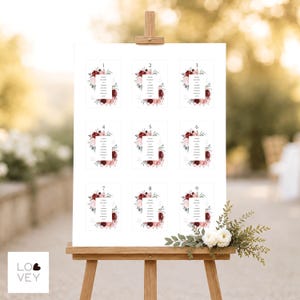 May include: A white wedding seating chart sign displayed on a wooden easel, featuring nine rectangular sections numbered one through nine. Each section includes a list of names framed by watercolor illustrations of deep red and pink flowers.