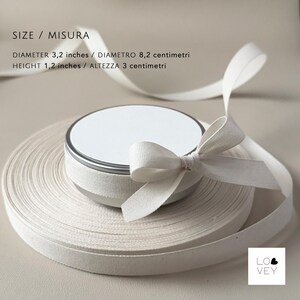 May include: A small, round, white container with a silver rim and a matching lid, sitting atop a roll of off-white ribbon. The container is tied with a white bow. The diameter is 3.2 inches, and the height is 1.2 inches.