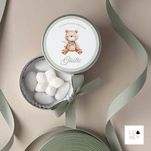 May include: A small, open tin filled with white candies and a matching lid. The lid features a watercolor illustration of a teddy bear with a green bow, the text "EccoMi Sono Arrivato" and the name "Giulio". A green ribbon is draped around the tin.