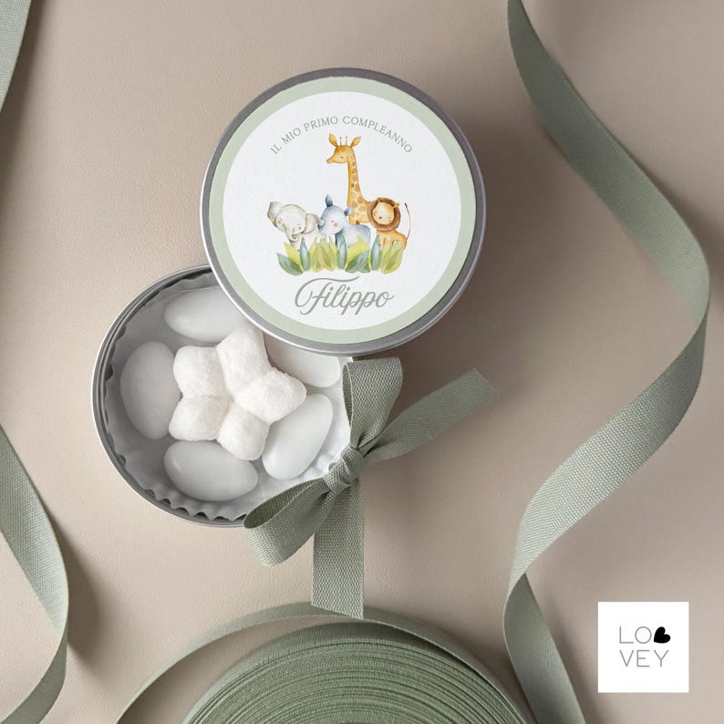 May include: A round, silver tin with a light green rim and lid, featuring a watercolor illustration of a giraffe, elephant, rhino, and lion. The text "IL MIO PRIMO COMPLEANNO" and "Filippo" are printed on the lid. The tin is open, revealing white candies.