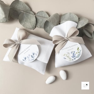 Könnte beinhalten: Zwei weiße Stoff-Gastgeschenktüten, gebunden mit beigen Schleifen. Jede Tüte hat ein rundes Etikett mit Namen und Datum, eines mit blauen Blättern und das andere mit blauen Blumen. Zwei weiße Mandeln liegen davor.
