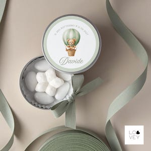 May include: A round, silver tin with a green and white label featuring a teddy bear in a hot air balloon. The tin is open, revealing white candies. The text "Davide" is written on the label. A green ribbon is draped around the tin.