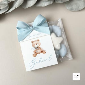 Può includere: Una scatola regalo con un fiocco azzurro e un'illustrazione di un orsacchiotto. La scatola ha il nome "Gabriel" e la parola "Benvenuto" stampati sopra. Accanto alla scatola c'è un sacchetto trasparente con caramelle blu e bianche.